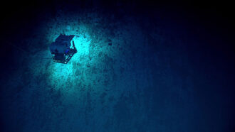 A photo of a remotely operated vehicle exploring the Mariana Trench. It appears as a cube hovering over a lighter blue patch surrounded by darkness.