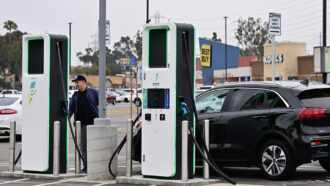 A photo of a man at an electric vehicle charging station with a black car parked next to him.