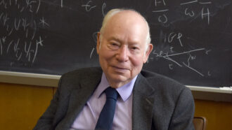 Steven Weinberg sitting in front of a chalkboard covered in equations